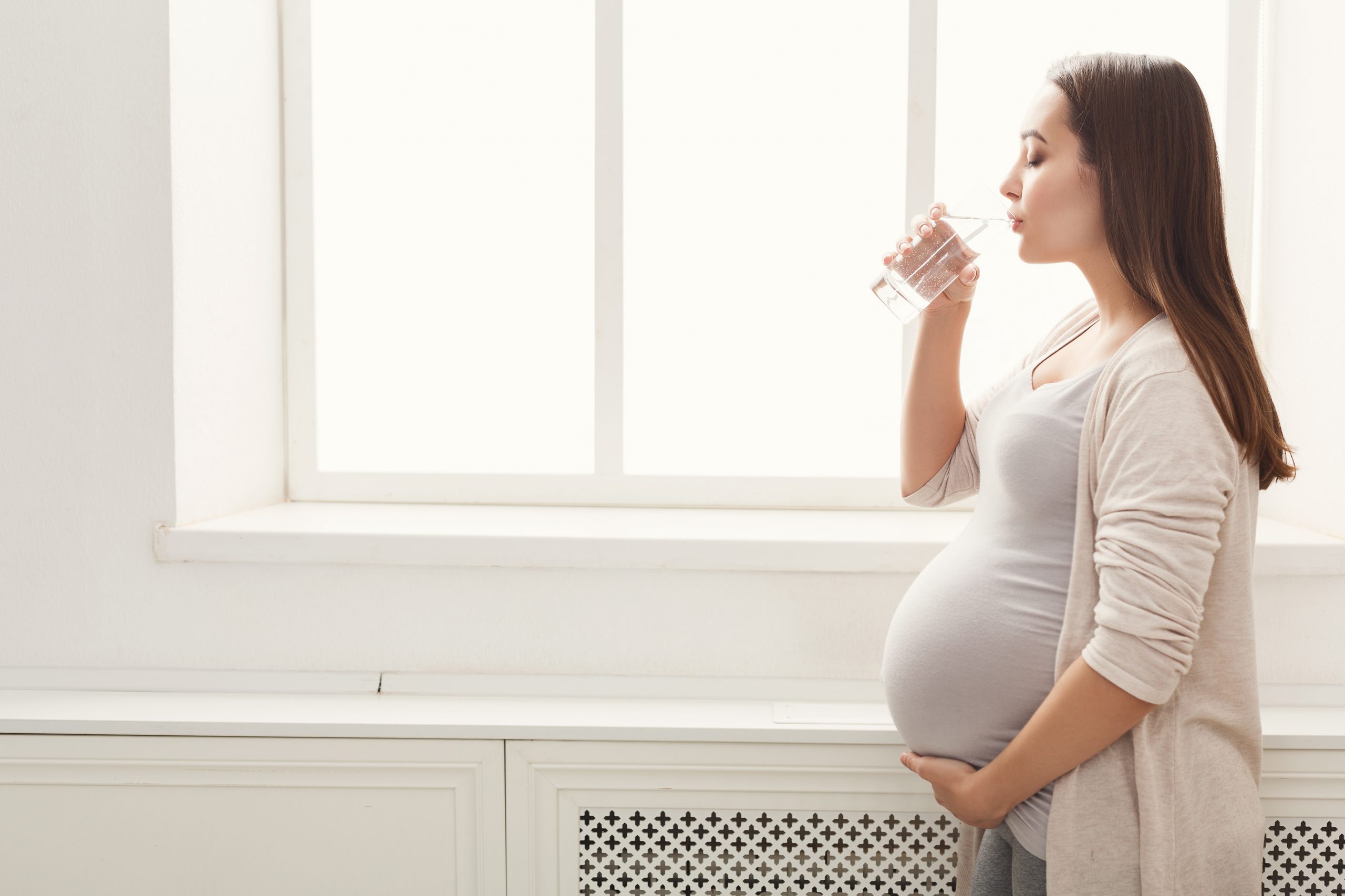 mulher grávida, segurando a barriga e tomando um copo de água enquanto observa a janela